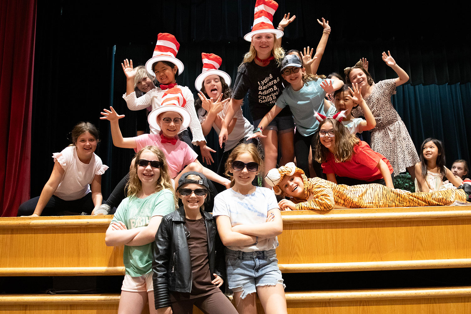 A group of children in playful costumes pose on a stage with red and white hats and sunglasses.