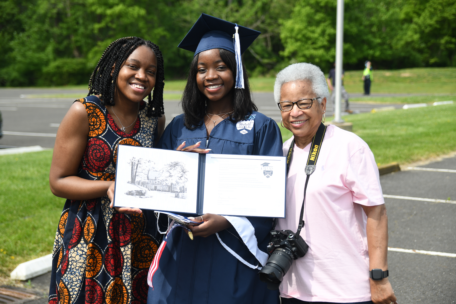 young woman graduating college and posing in cap and gown with diploma, accompanied by two guests