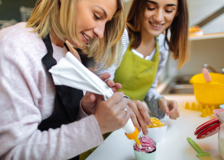 Woman piping icing on a cupcake with friend helping in background.