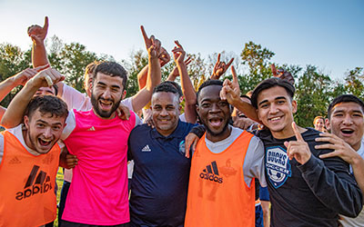 Six men in soccer apparel smile and celebrate together outdoors, with several raising their hands.
