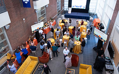Overhead view of a busy formal reception in a brick-and-glass atrium, with guests gathered around gold-covered cocktail tables and buffet stations.