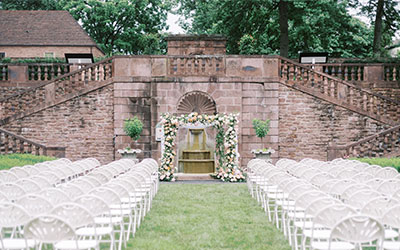 Outdoor wedding ceremony setup with white chairs lining a grassy aisle toward a flower-decorated stone altar.