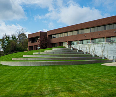 Outdoor amphitheater with curved concrete seating tiers, a grassy lawn, and a modern brick campus building behind it.