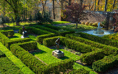 Formal garden with geometric boxwood hedges, two pedestal sculptures, and a stone fountain surrounded by autumn trees.