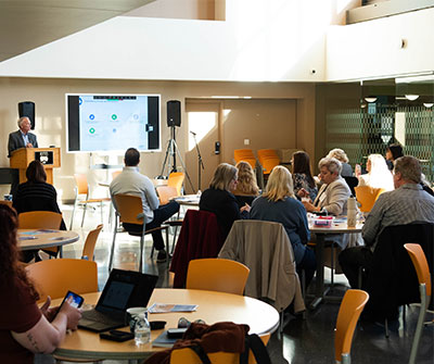 A speaker presents to an audience seated at round tables in a conference room.
