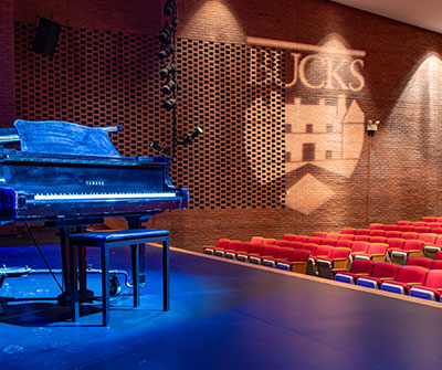 A grand piano on a blue-lit theater stage facing rows of red seats, with a brick wall and “Bucks” logo in the background.