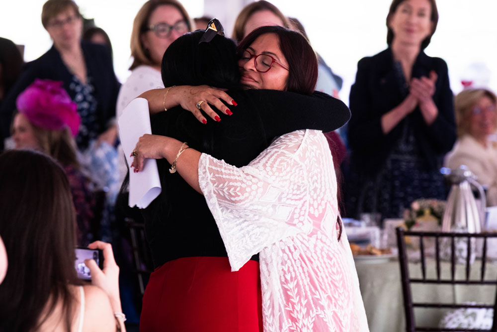 Recipient Kassandra Castillo and her mother embrace warmly in a room filled with people. The atmosphere is joyful, with onlookers clapping. Tables with floral decorations are visible.