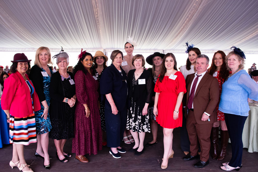 A group of attendees, mostly women, pose under a white canopy. They wear formal attire with hats, smiling warmly, suggesting a festive event.