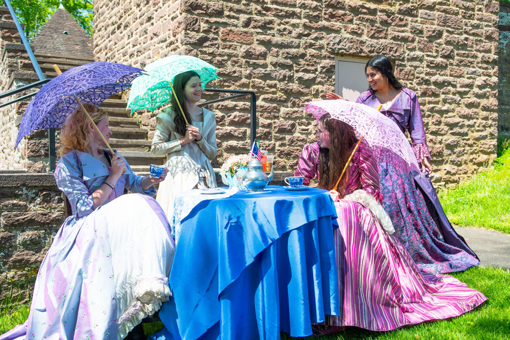 Four women in vibrant, Victorian-style dresses enjoy a sunny outdoor tea party. They hold parasols, with a blue-clothed table and teacups nearby.