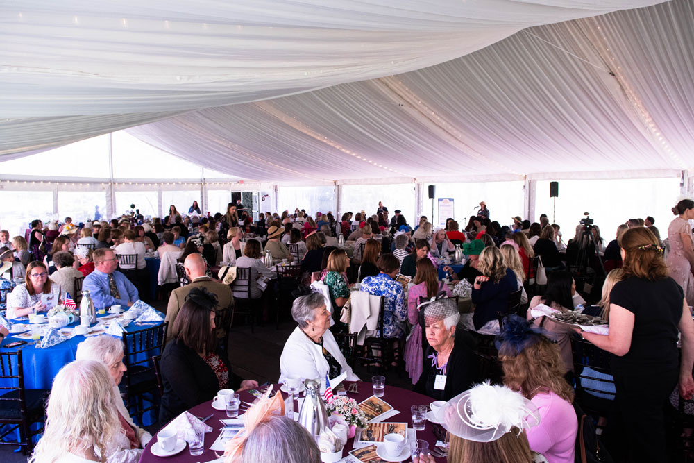 A large group of people seated at round tables in a spacious, draped tent. Many wear hats, creating a vibrant and festive atmosphere.