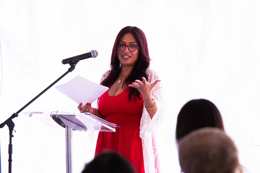 Recipient Kassandra Castillo in a red dress and glasses speaking passionately at a podium with a microphone, holding papers. Audience in foreground, blurred.