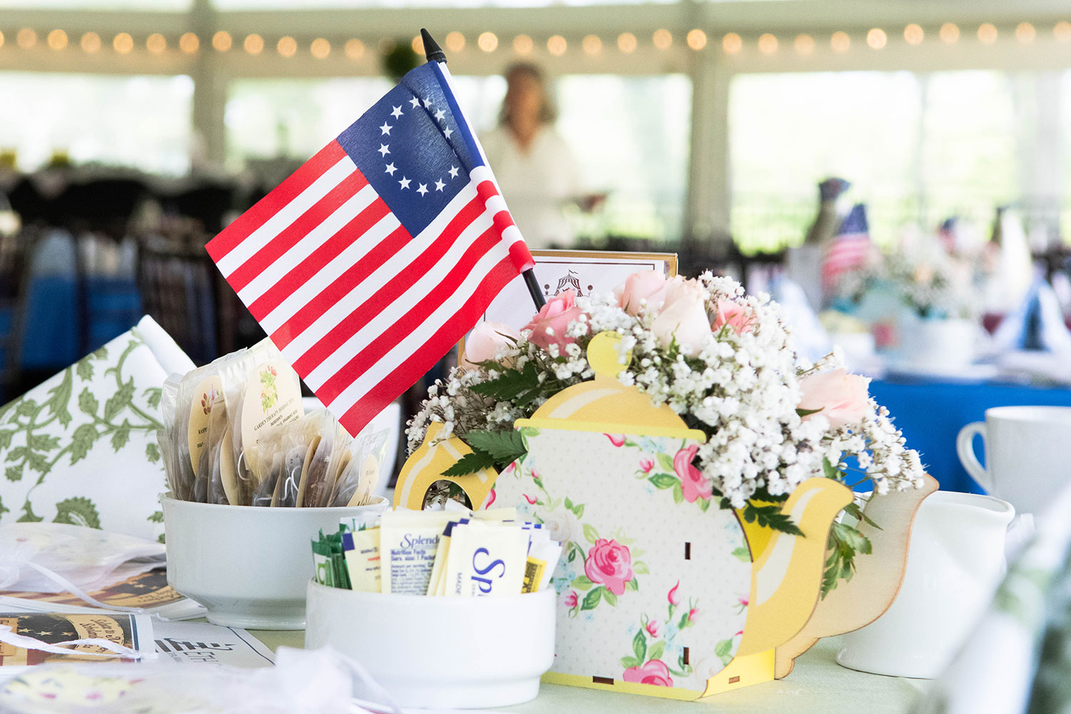 A Betsy Ross flag decorates a table with a floral centerpiece in a teapot-shaped holder. Teabags and sweeteners are in small bowls nearby, creating an elegant, inviting atmosphere.