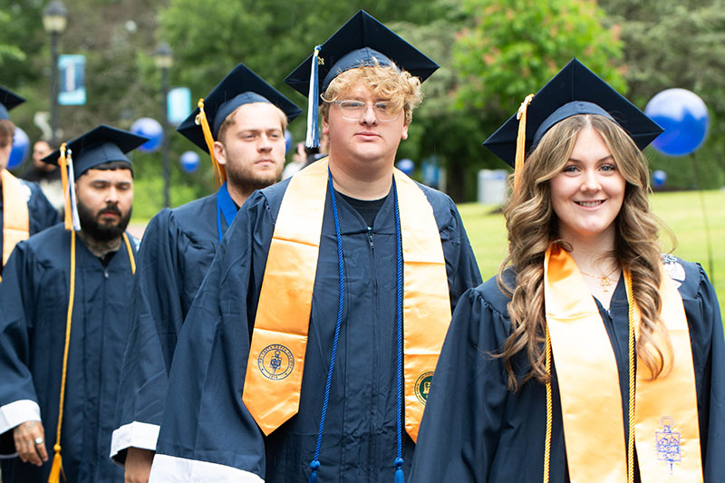 Graduates in caps and gowns walk in a procession outdoors, smiling. They wear yellow stoles, with blue and white tassels, conveying joy and achievement.