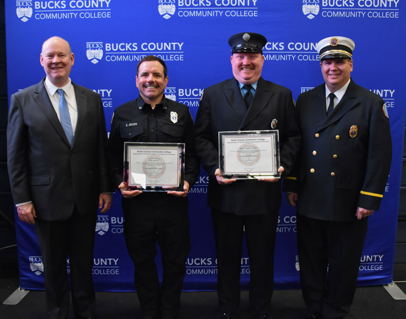 Four men in formal attire, with two in uniforms holding framed awards, stand smiling in front of a Bucks County Community College backdrop.