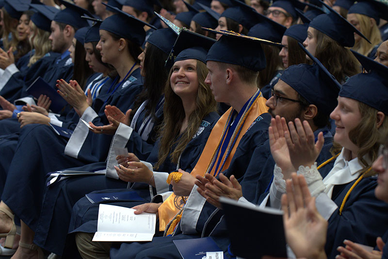 A group of graduates in blue caps and gowns sit in rows, smiling and clapping. The atmosphere is joyful and celebratory.
