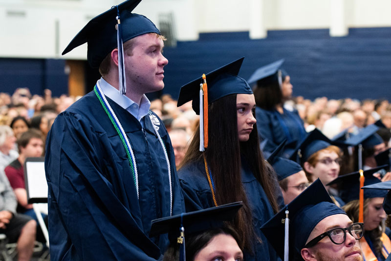 Bucks graduates in navy caps and gowns stand in a packed auditorium during a ceremony. They look focused and proud, surrounded by seated peers and a large audience.