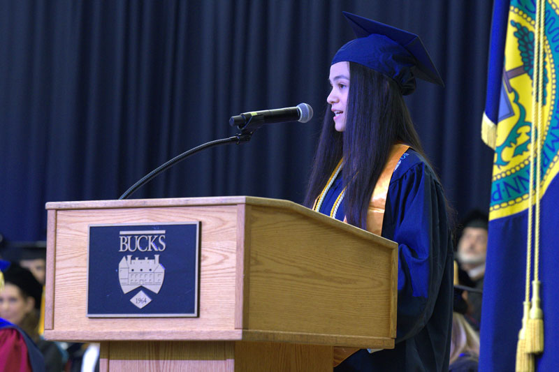 Student speaker, Lia Peralta Joa, in cap and gown speaks confidently at a podium labeled 