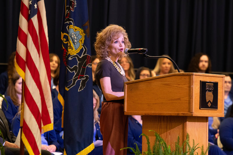 Teresa McMonagle, CRNP, RN speaks at a podium with a microphone during a formal event, flanked by the U.S. and Pennsylvania flags. An attentive audience sits behind her.