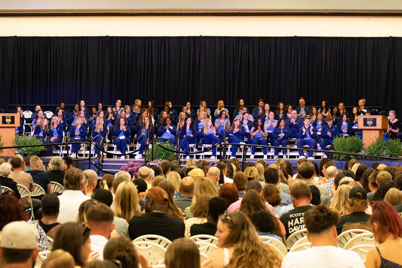 Audience seated in a hall watching a AD Nursing pinning ceremony. A group in blue robes sits on stage, applauding, with podiums flanking them.