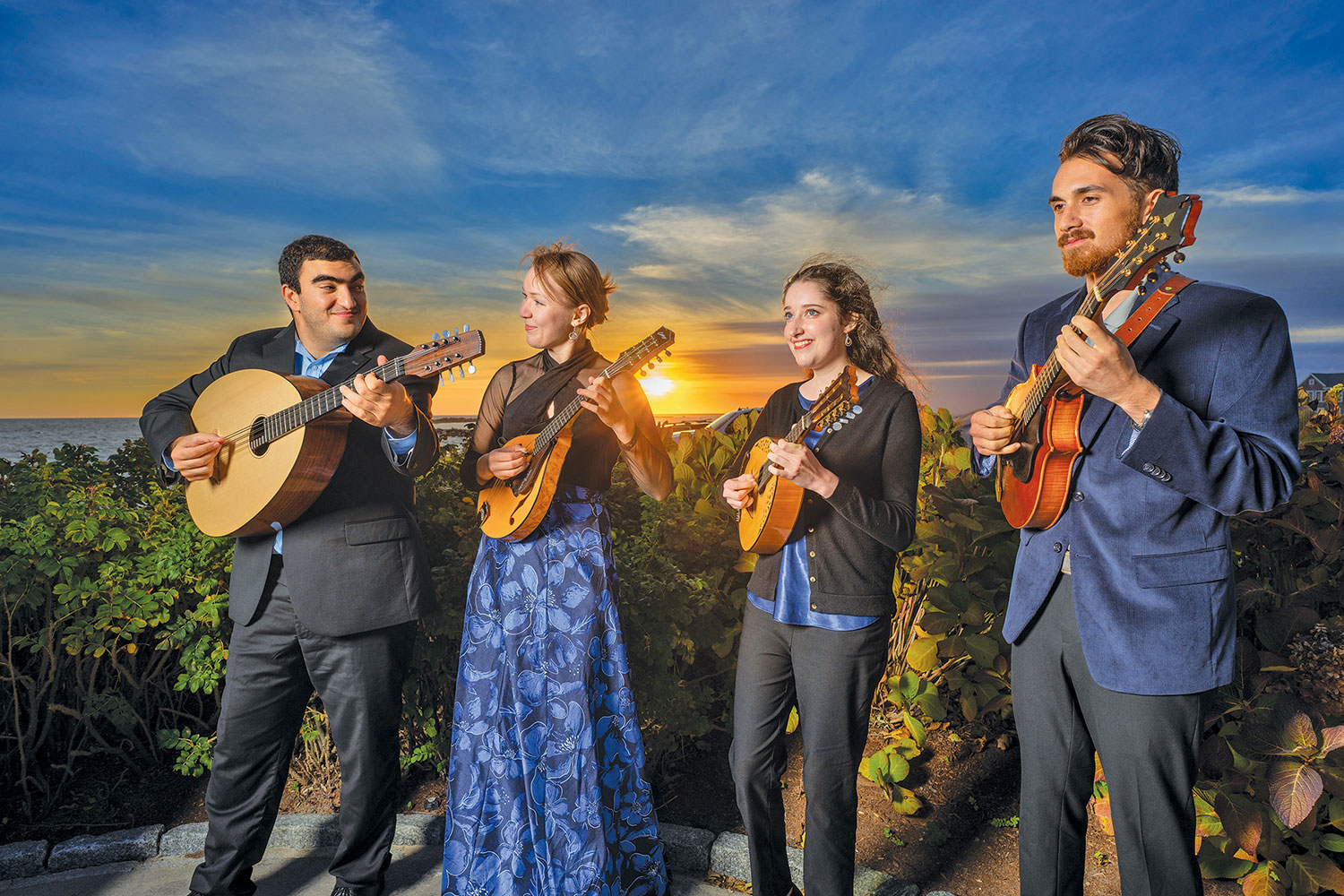 The Atlantic Mandolin Quartet in formal attire play string instruments against a sunset backdrop by the ocean. The scene conveys joy and harmony.