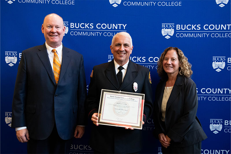 A firefighter in uniform holds a certificate, flanked by a man in a suit and a woman in a blazer, all smiling.