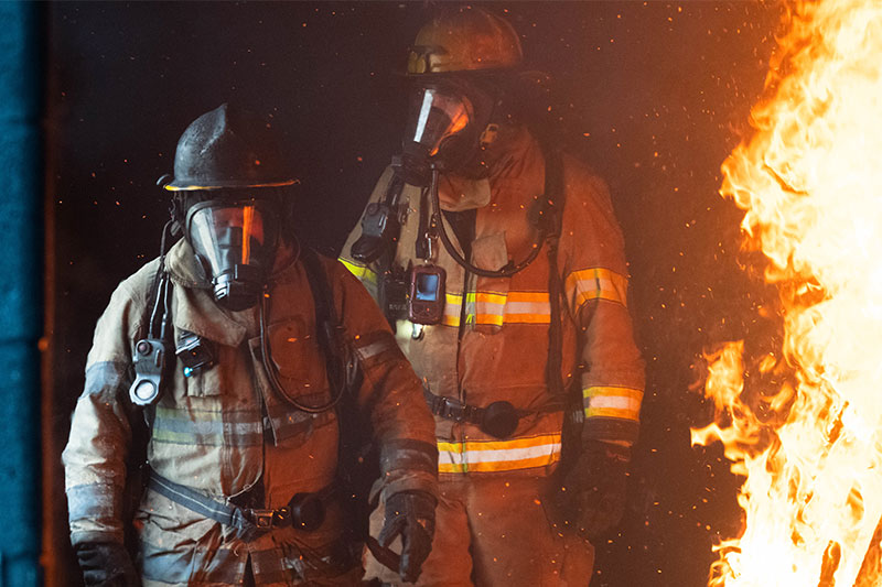 Two firefighters in protective gear stand before a large fire, ready for action in a dark environment.