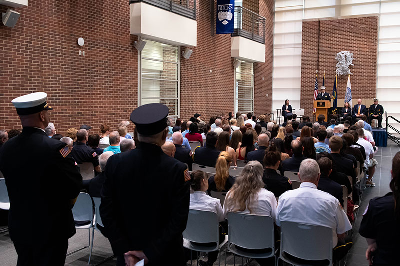 A graduation ceremony at Bucks County Community College Fire School with graduates and families in attendance, featuring a speaker at a podium surrounded by flags.