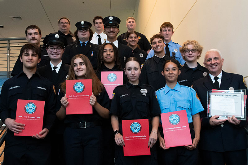 Group photo of Bucks County Community College Fire School graduates holding red certificate folders, arranged on a staircase in uniforms.