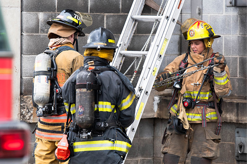 Three firefighters in gear collaborate near a ladder against a concrete wall, with one managing a chain while the others look on.