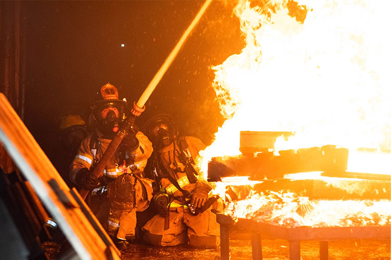 Firefighters in protective gear are extinguishing flames from burning wooden pallets with a water hose, amidst a smoky, dark background.