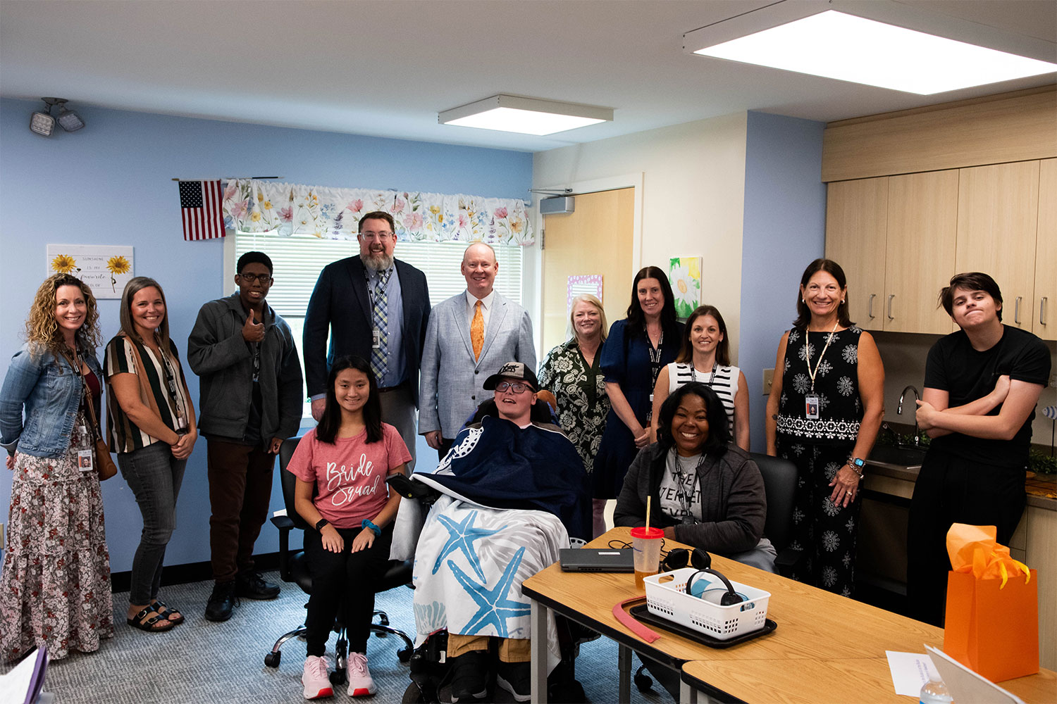 Group of 13 people posing in an office, including a person seated in a wheelchair.
