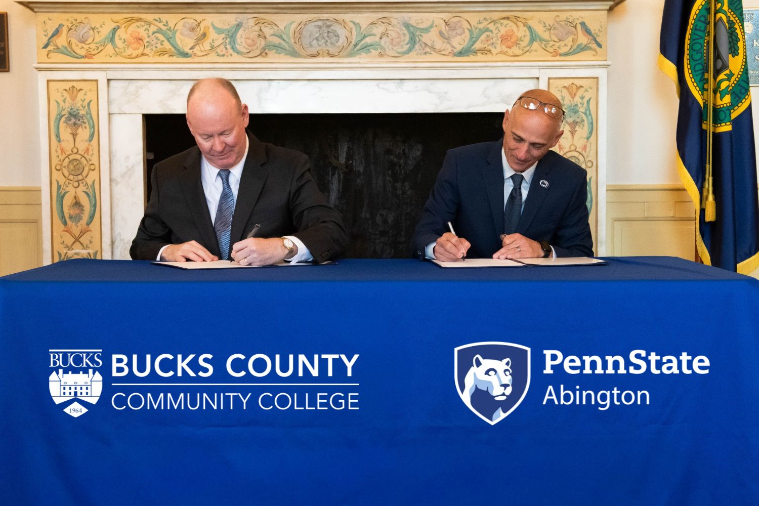 The president of Bucks County Community College and the chancellor of Penn State Abington sign documents at a blue-draped table.