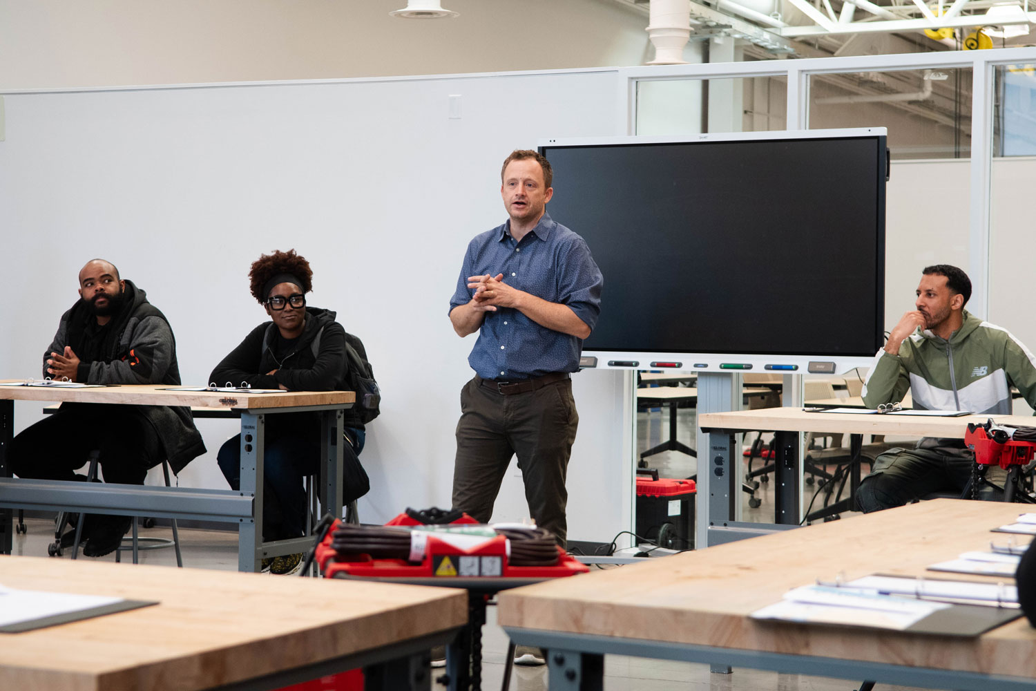 A man stands speaking in front of a large screen in a classroom, engaging three seated people at tables.