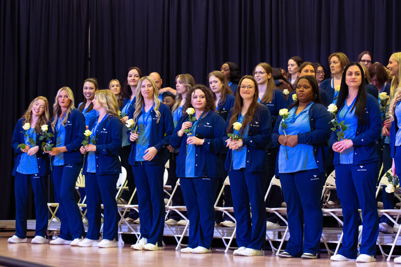 A group of nursing graduates in blue uniforms stand proudly, holding white roses. They are on a stage, with an audience in the background, conveying achievement.