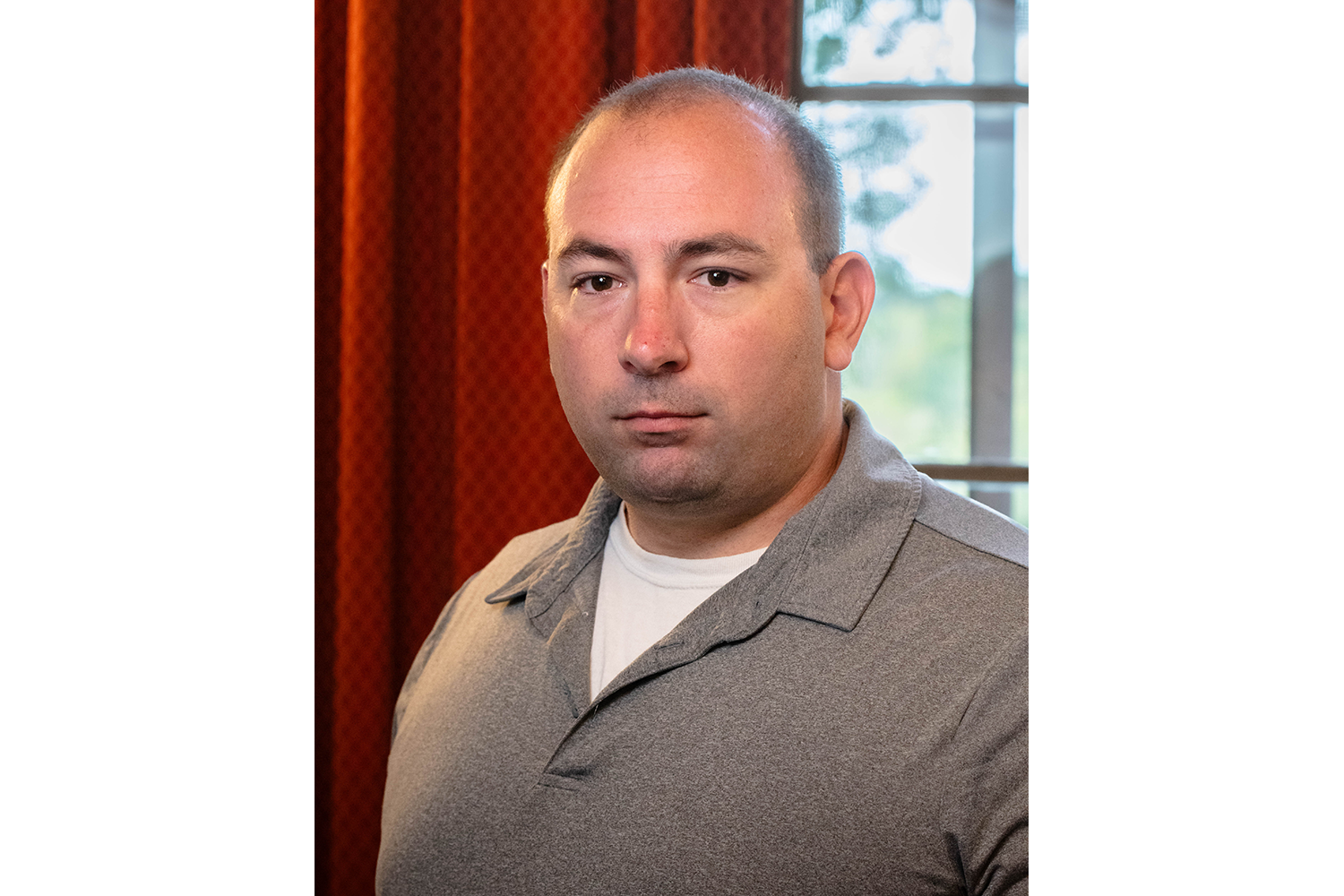 Close-up portrait of a Ryan Ross in a gray collared shirt, standing indoors beside a red curtain and window.